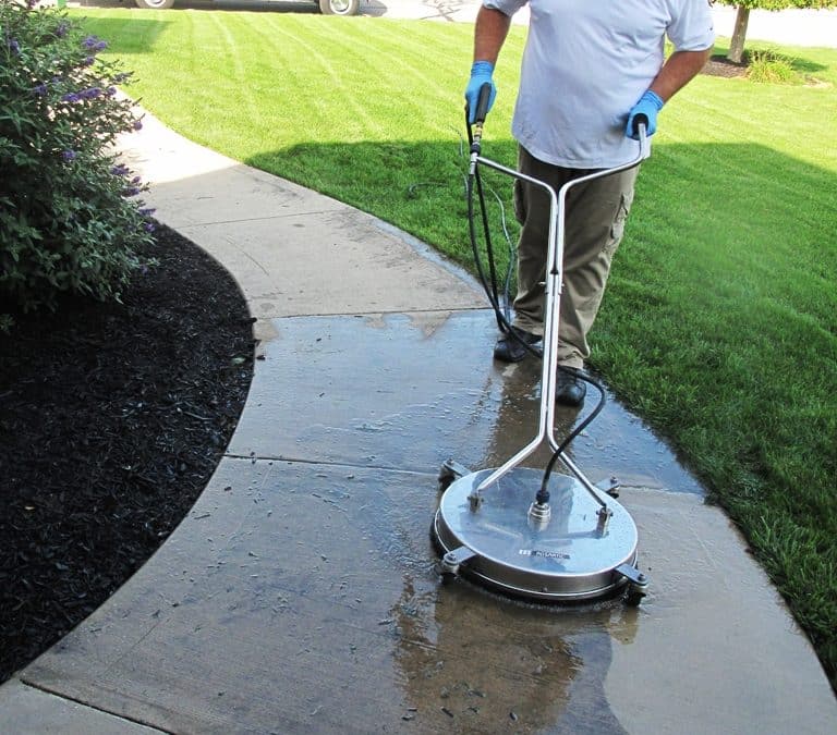 A technician uses a surface cleaner to wash a concrete sidewalk, showing a clear contrast between cleaned and uncleaned sections, highlighting professional concrete cleaning services.