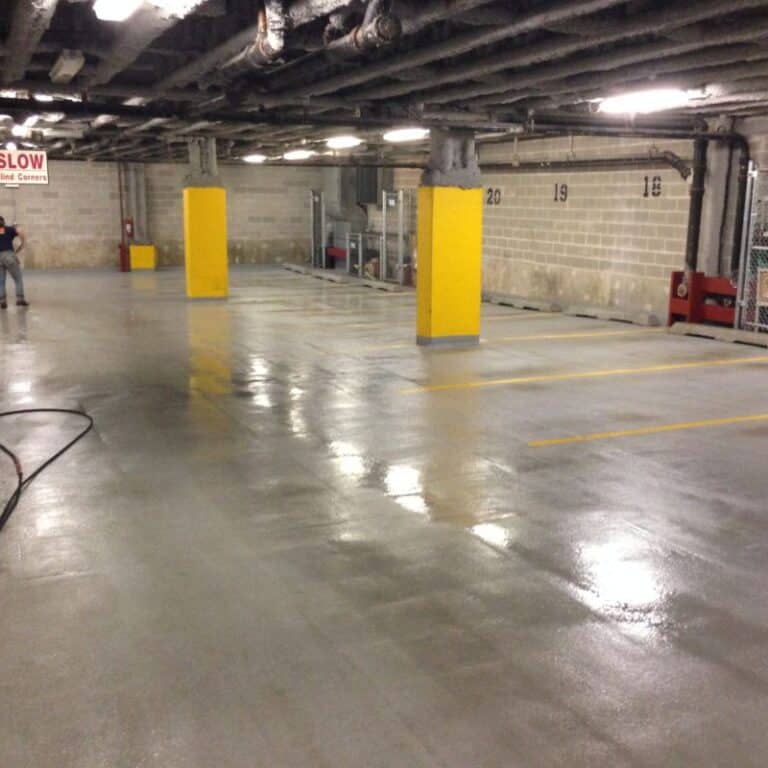 A clean, reflective concrete floor in an underground parking garage with yellow-painted columns and visible lighting, demonstrating the effectiveness of commercial pressure washing services.
