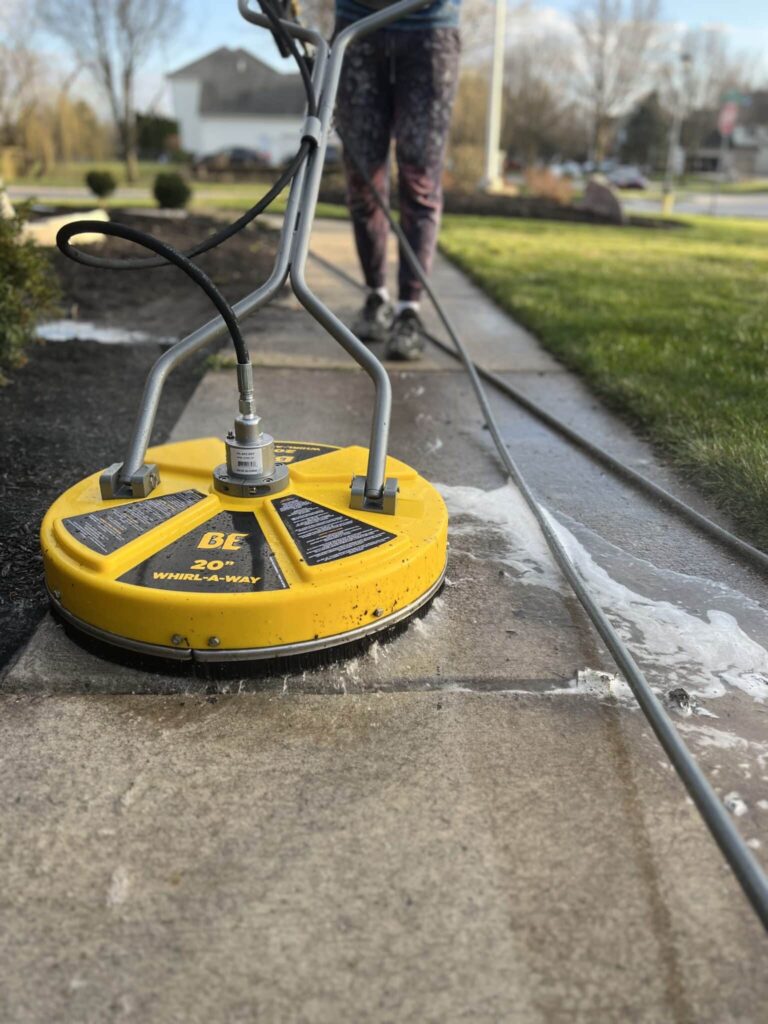 A technician uses a yellow 20-inch surface cleaner to wash a concrete sidewalk, creating a visible contrast between cleaned and uncleaned sections, demonstrating effective pressure washing in Holland, OH.