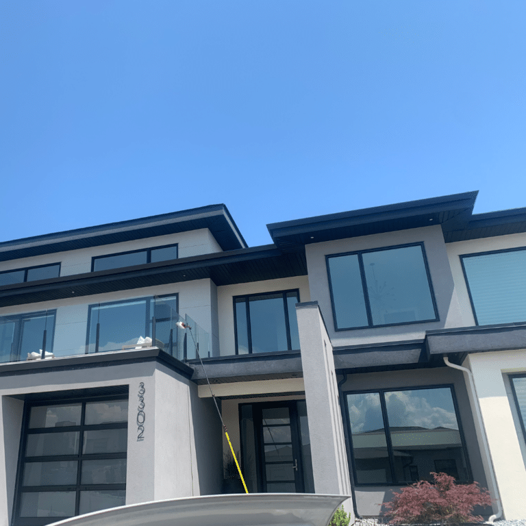 A long water-fed pole reaches up to clean large windows on a two-story modern home under a clear blue sky, showcasing precision and safety in exterior window maintenance.