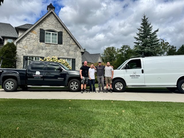 Four individuals stand together on a driveway in front of a stone house, flanked by a black pickup truck and a white van. The scene features a well-maintained lawn and overcast skies.