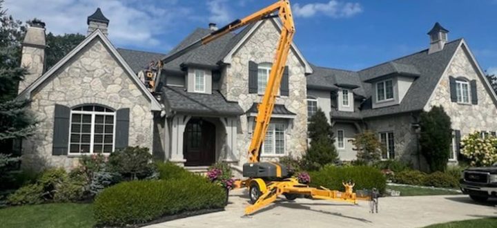 A large stone house with dark shutters and a gray roof is shown under a bright blue sky. A yellow boom lift is positioned on the driveway, indicating exterior maintenance or painting work. The property features well-manicured landscaping and a clean concrete driveway.